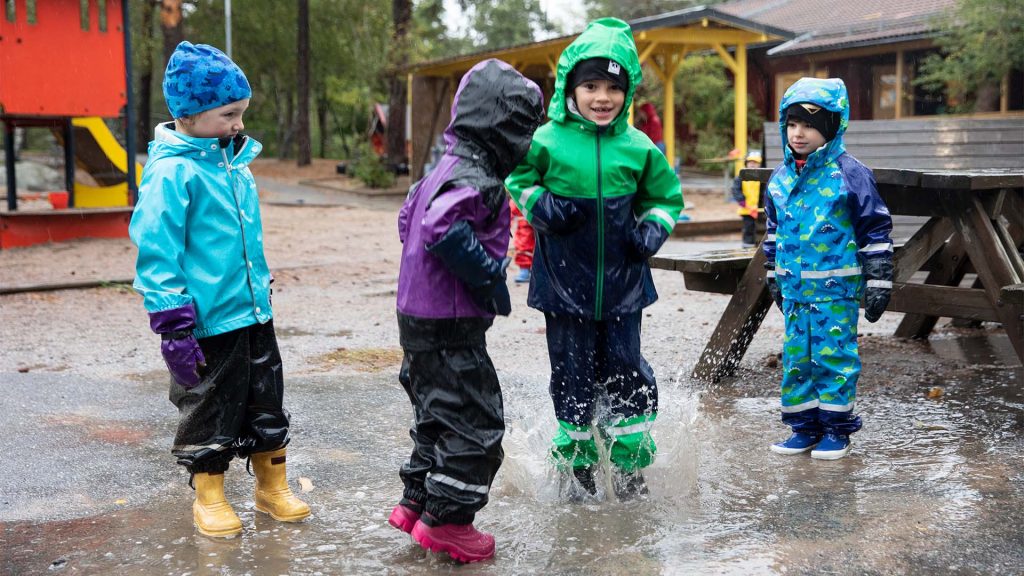 Fyra barn är i skolgården och har regnkläder på sig, de hoppar i en stor vattenpöl