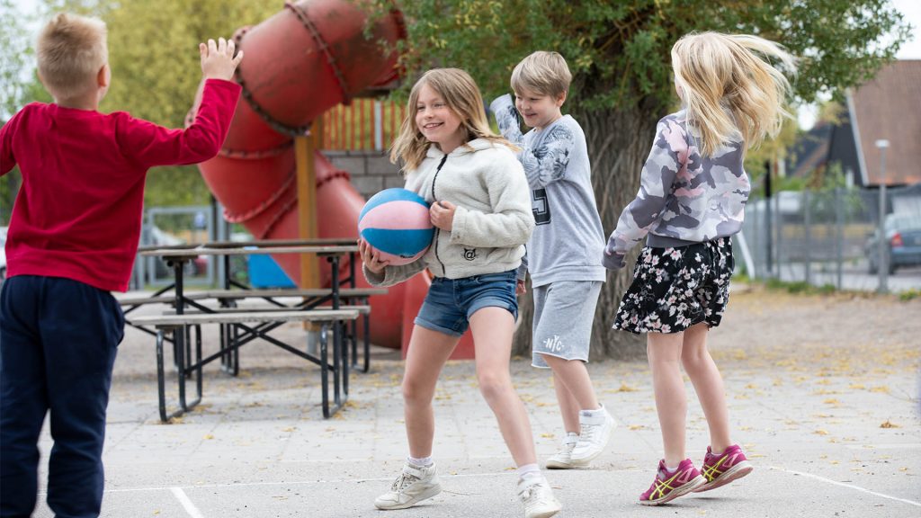 Barn leker vid en skolgård, en flicka håller i en basketboll och en pojke hoppar