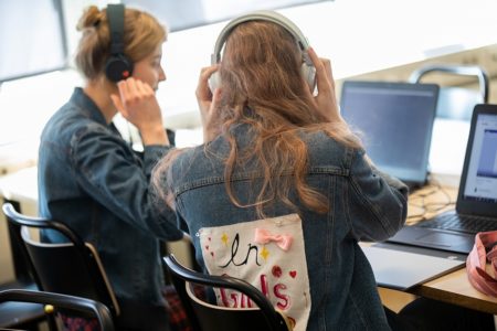 Två elever sitter bredvid varandra i klassrummet, de har båda på sig hörlurar och tittar på en datorskärm
