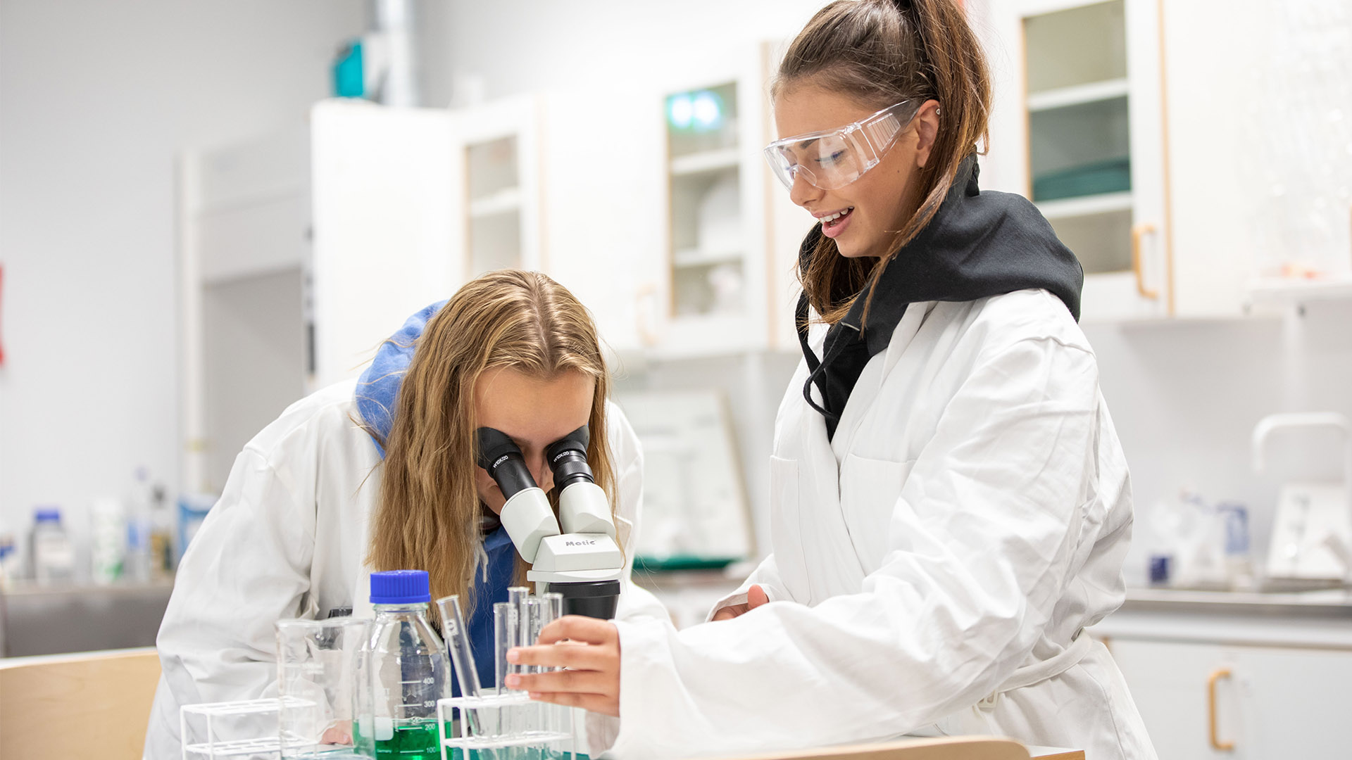 Två elever i vita labbrockar, en av de ser in i ett mikroskop medan den andra har skyddsglasögon och ser på