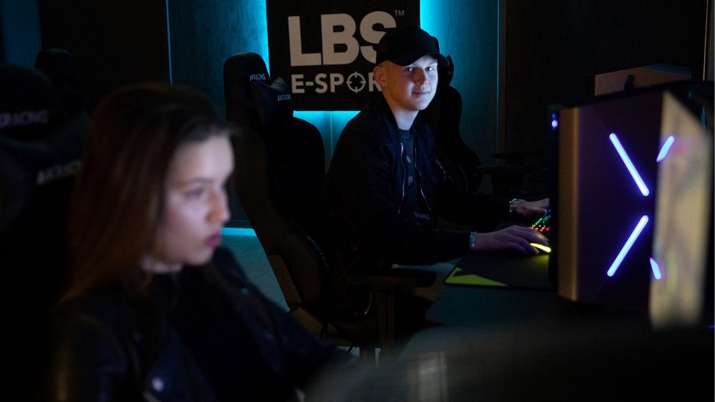 Två personer sitter i ett mörkt rum och spelar datorspel, på väggen står en skylt med "LBS E-Sport"