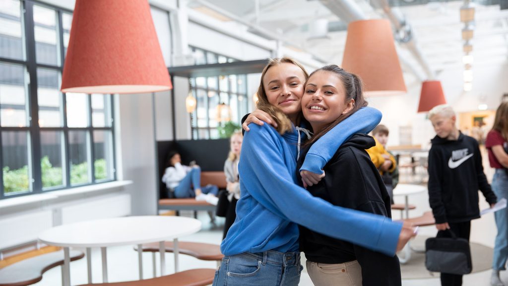Två tjejer håller om varandra och ler framför kameran i en kafeteria-miljö