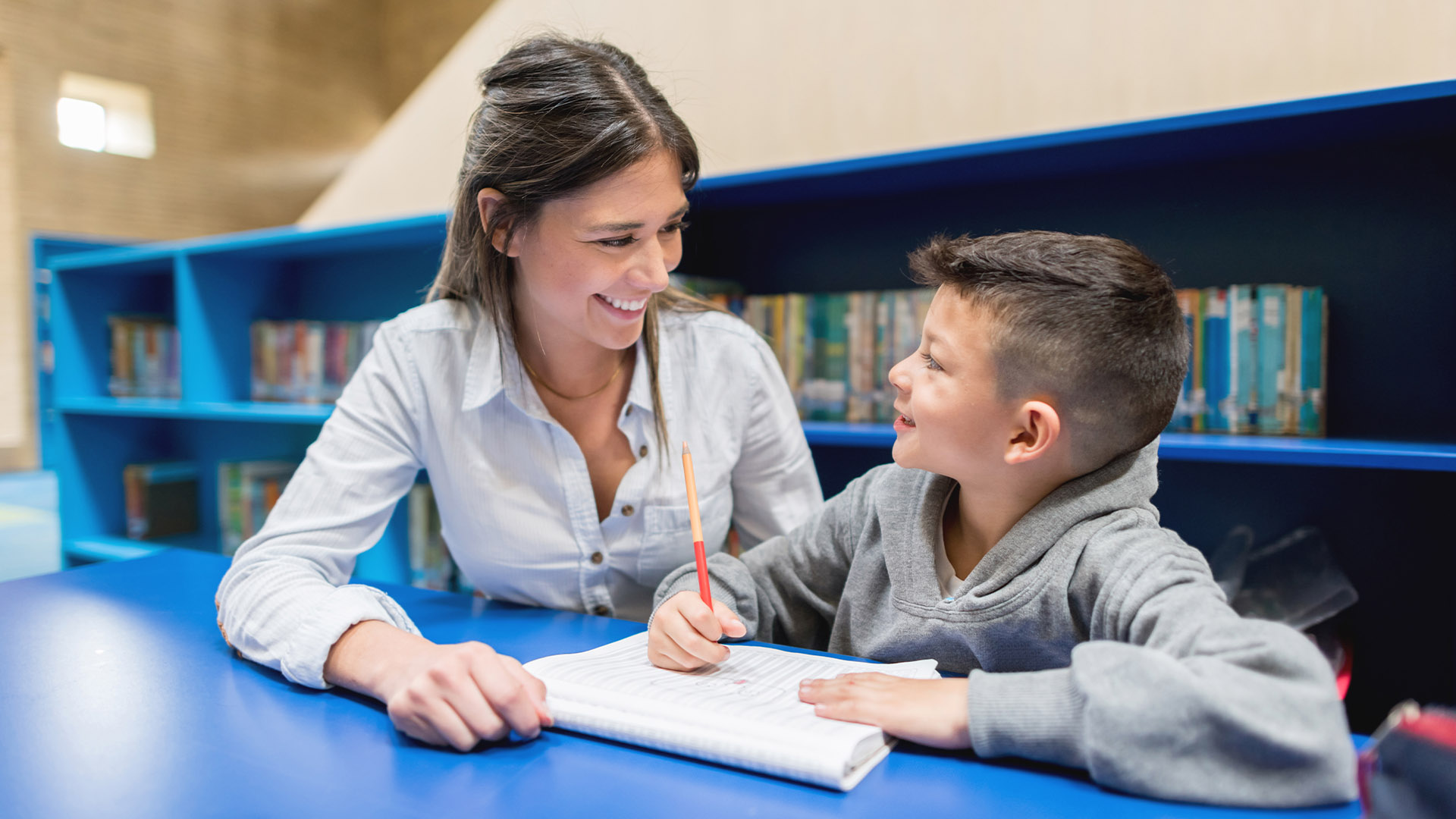 Lärare och ung elev sitter i ett bibliotek, den unga pojken skriver i hans anteckningsbok och läraren ler mot honom.