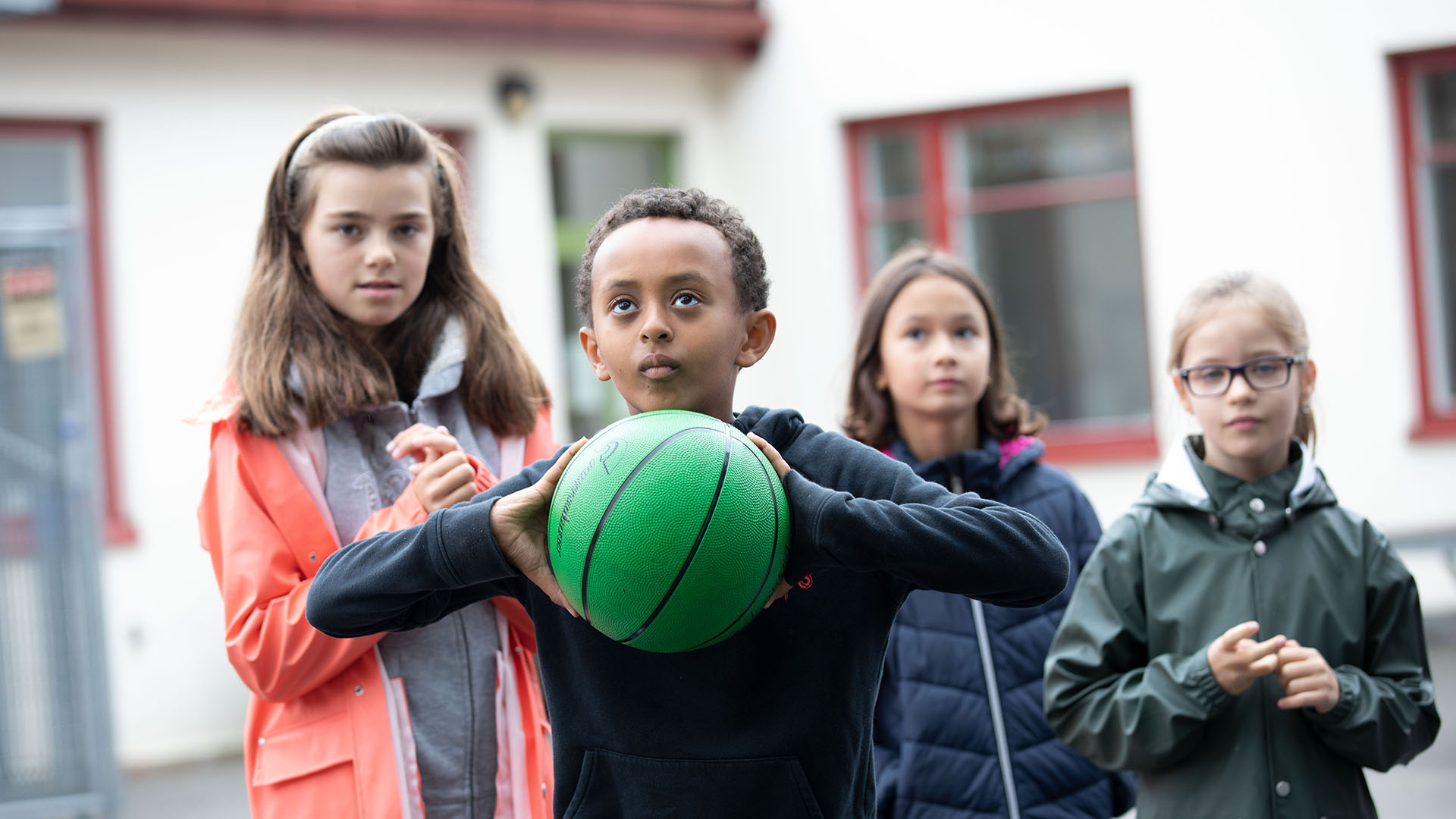 Pojke i centrum som fokuserar på kasta sin gröna basketboll, bakom honom är tre barn som tittar på