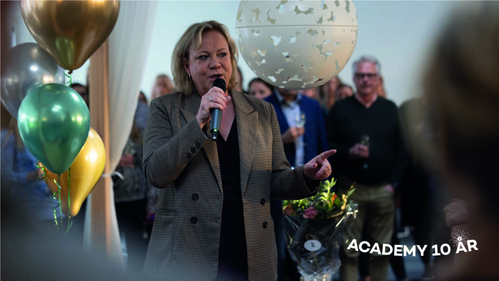 Lisa Oldmark står med en mikrofon i handen och håller ett tal. I bakgrunden syns människor och ballonger. På bilden står det Academy 10 år.