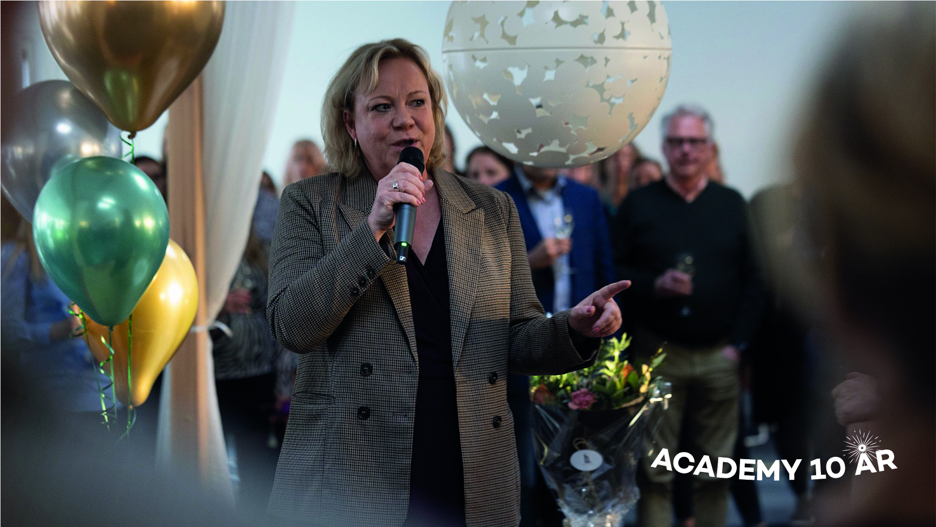 Lisa Oldmark står med en mikrofon i handen och håller ett tal. I bakgrunden syns människor och ballonger. På bilden står det Academy 10 år.